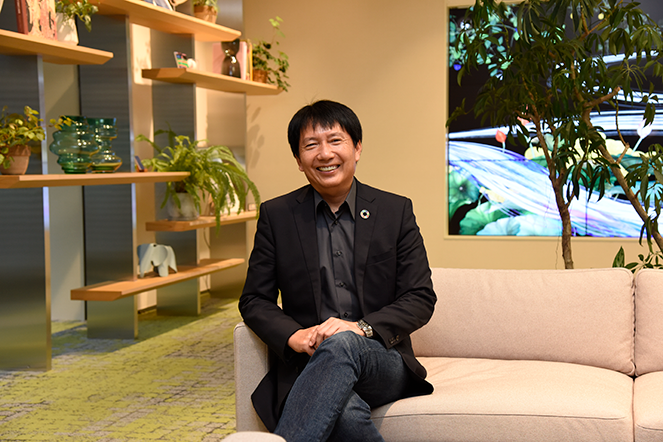 A business professional sits on a light-colored sofa in a modern office lounge, smiling at the camera. The background features shelves with plants and decorative items as well as greenery and a large screen displaying vibrant imagery.