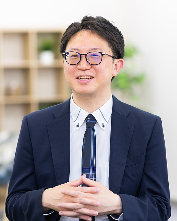 A person wearing a navy suit, white shirt, and striped tie, with hands clasped, standing in a modern office setting with shelves and greenery in the background.