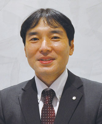A person wearing a dark suit, white shirt, and patterned tie against a neutral background.