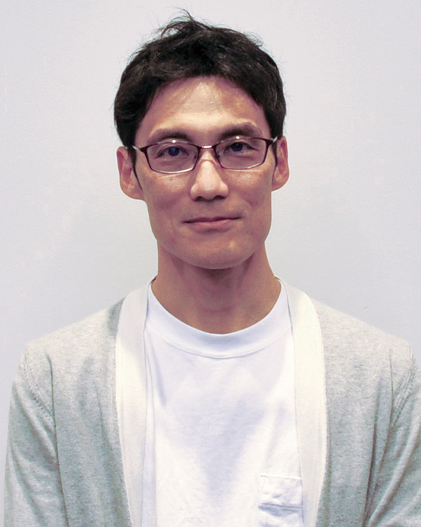 A portrait of a case study speaker from NRI, wearing glasses and a light sweater over a white shirt, standing against a plain background.