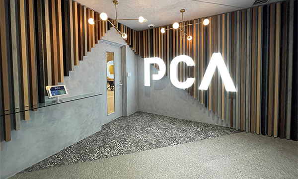 Modern office reception area featuring the illuminated PCA logo on a textured wall, stylish lighting fixtures, and a glass counter with a digital tablet display.