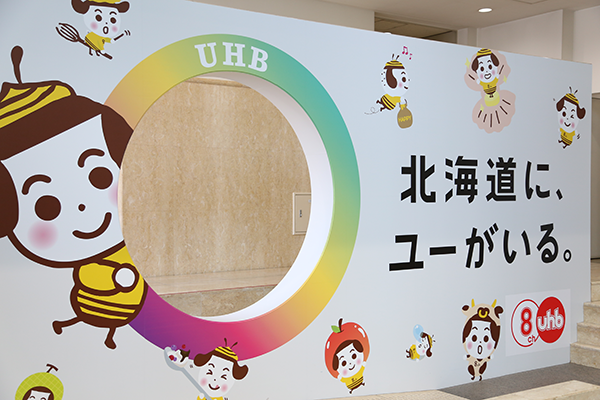 Photo of a colorful UHB Hokkaido Culture Broadcasting display board with cartoon characters, a circular cutout, and Japanese text in a public area.