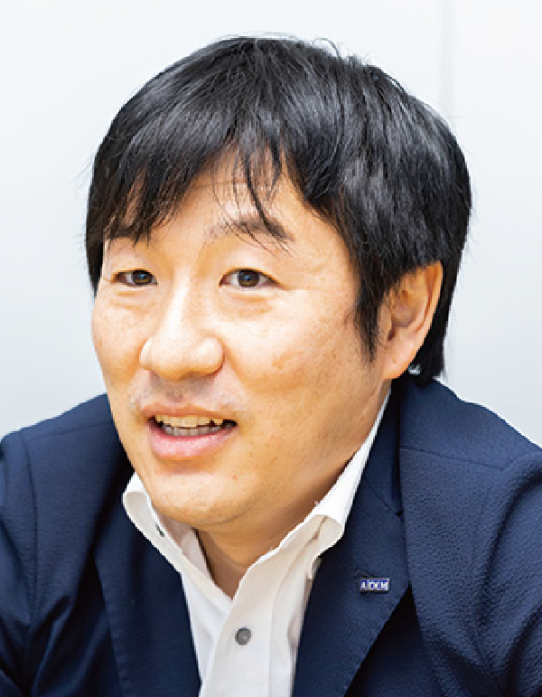 A businessman speaking, wearing a dark blazer with an AIDEM logo pin, and a white shirt.
