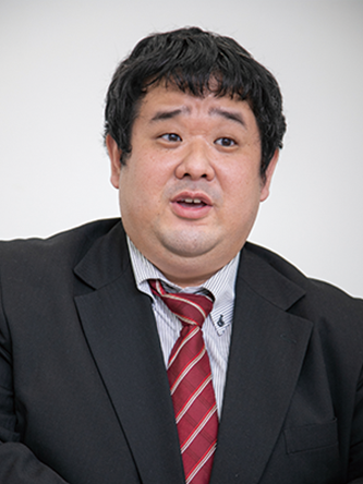 A man wearing a business suit and red striped tie, speaking or presenting against a plain background.