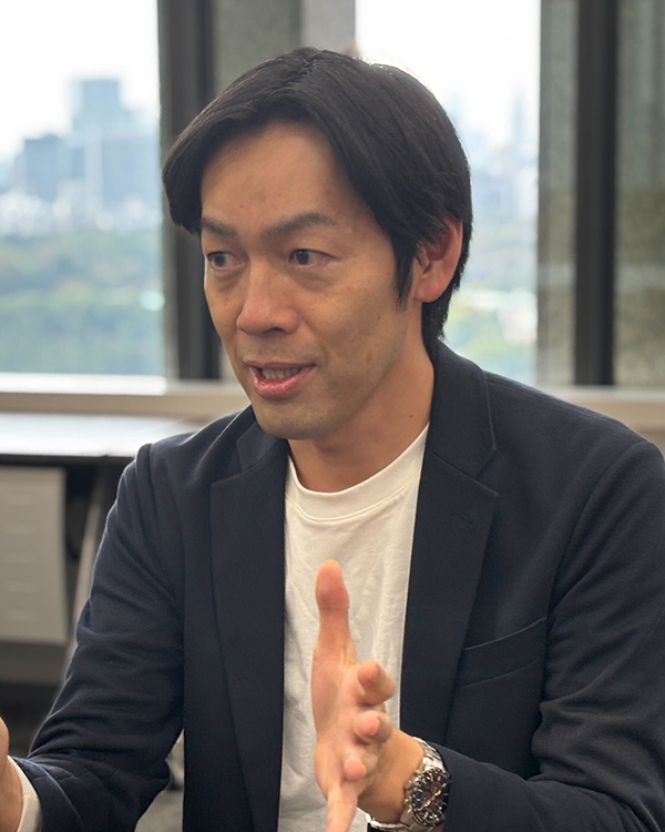 A person wearing a dark blazer and white shirt gesturing with hands, seated in an office with a cityscape visible through the window.
