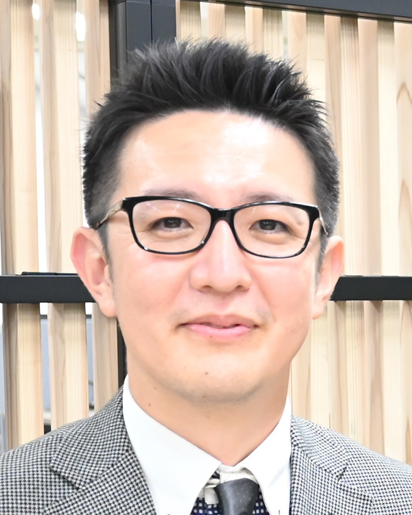 A person wearing a gray checkered suit, white shirt, and patterned tie, standing in front of a wooden slat background.