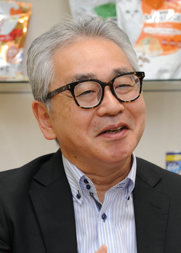 Portrait of a Nestlé executive wearing glasses and a suit, sitting indoors and speaking during an interview, with packaged products visible in the background.