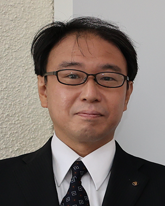 Professional portrait of a man wearing a suit, tie, and glasses, standing in front of a light-colored wall and looking at the camera.