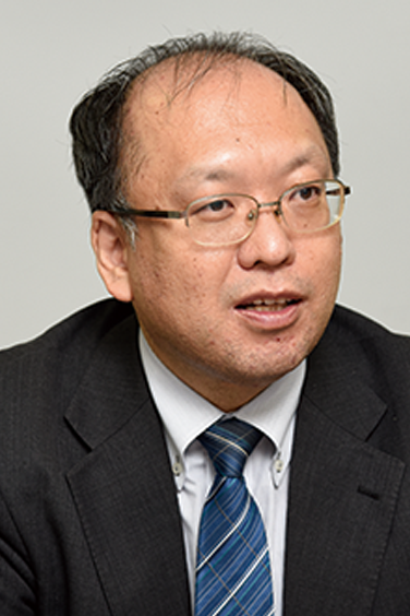 A businessman wearing a suit, tie, and glasses speaking or engaged in conversation. Portrait orientation with a neutral background.
