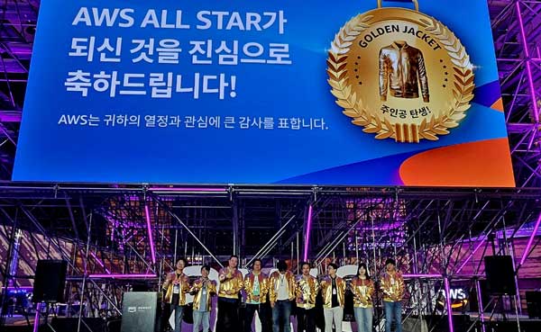 A group of people wearing golden jackets stand on a stage under a large screen displaying a congratulatory message in Korean for AWS All Star winners, with a "Golden Jacket" emblem.