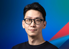 A portrait of a man with short black hair and glasses, wearing a dark shirt, posed against a vibrant blue and green gradient background with a geometric red and blue accent.