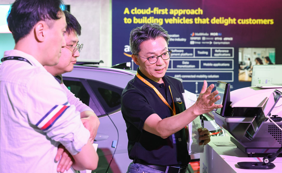 A person wearing an AWS shirt demonstrates technology to two individuals near a car, with a display about cloud-based vehicle solutions in the background.