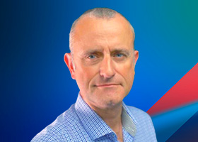 A professional headshot of a middle-aged man in a checkered shirt posing in front of a blue gradient background with red and teal accents.