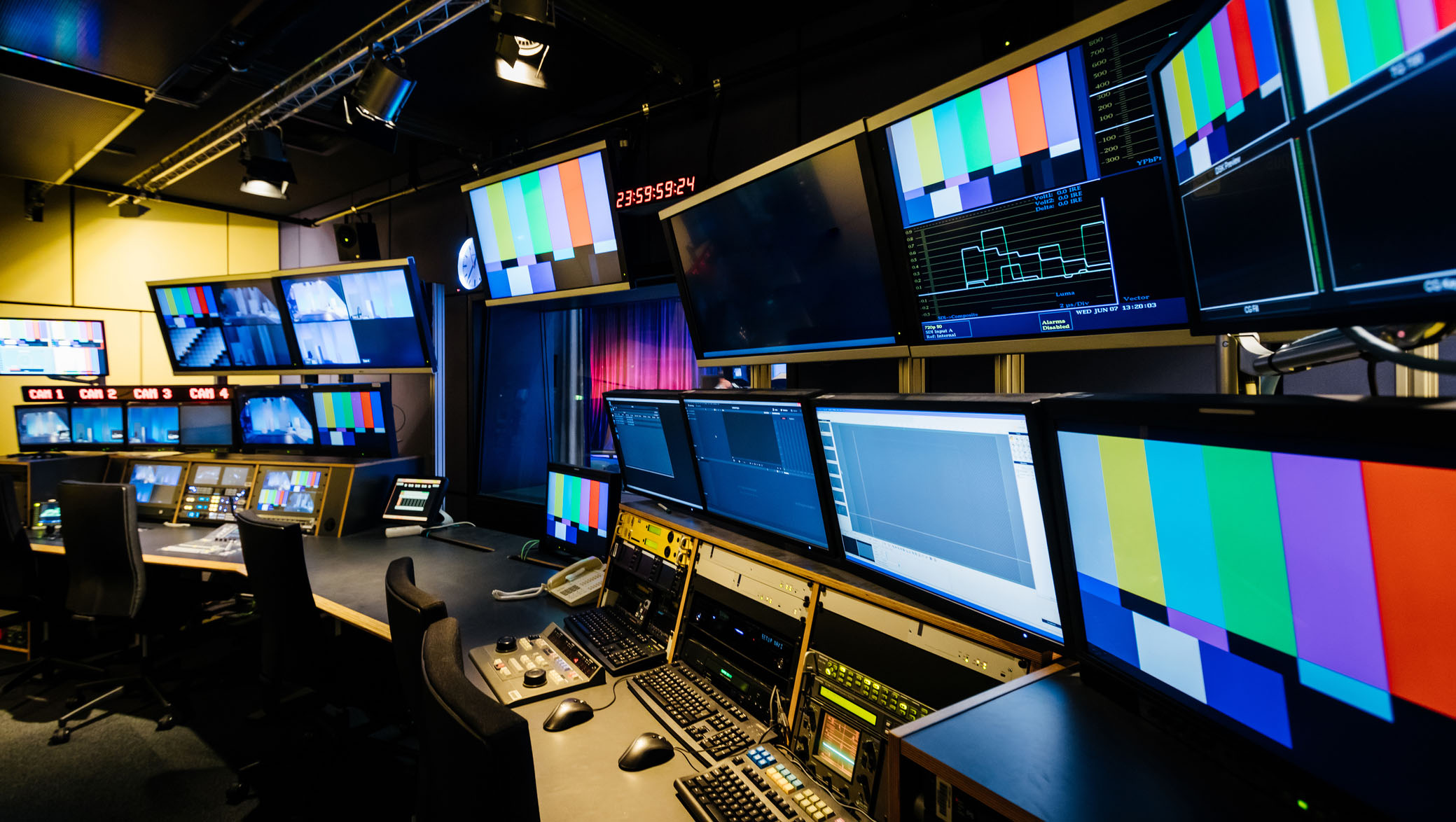 A modern university television video control room featuring multiple monitors displaying test patterns and camera feeds, audio and video equipment, and workstations for video production staff.