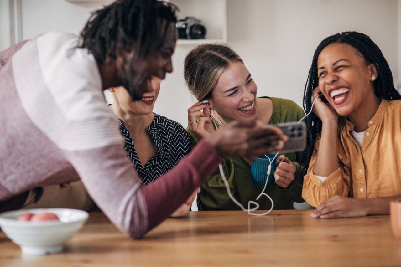 A group of friends laughing and enjoying while sharing earphones and watching something on a phone together at a table.
