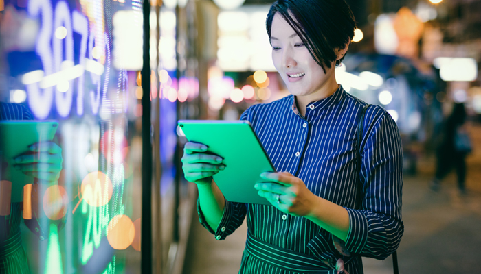 A woman stands in front of a brightly lit digital display in a city at night, using a tablet device. The scene suggests technology, security, and digital connectivity.