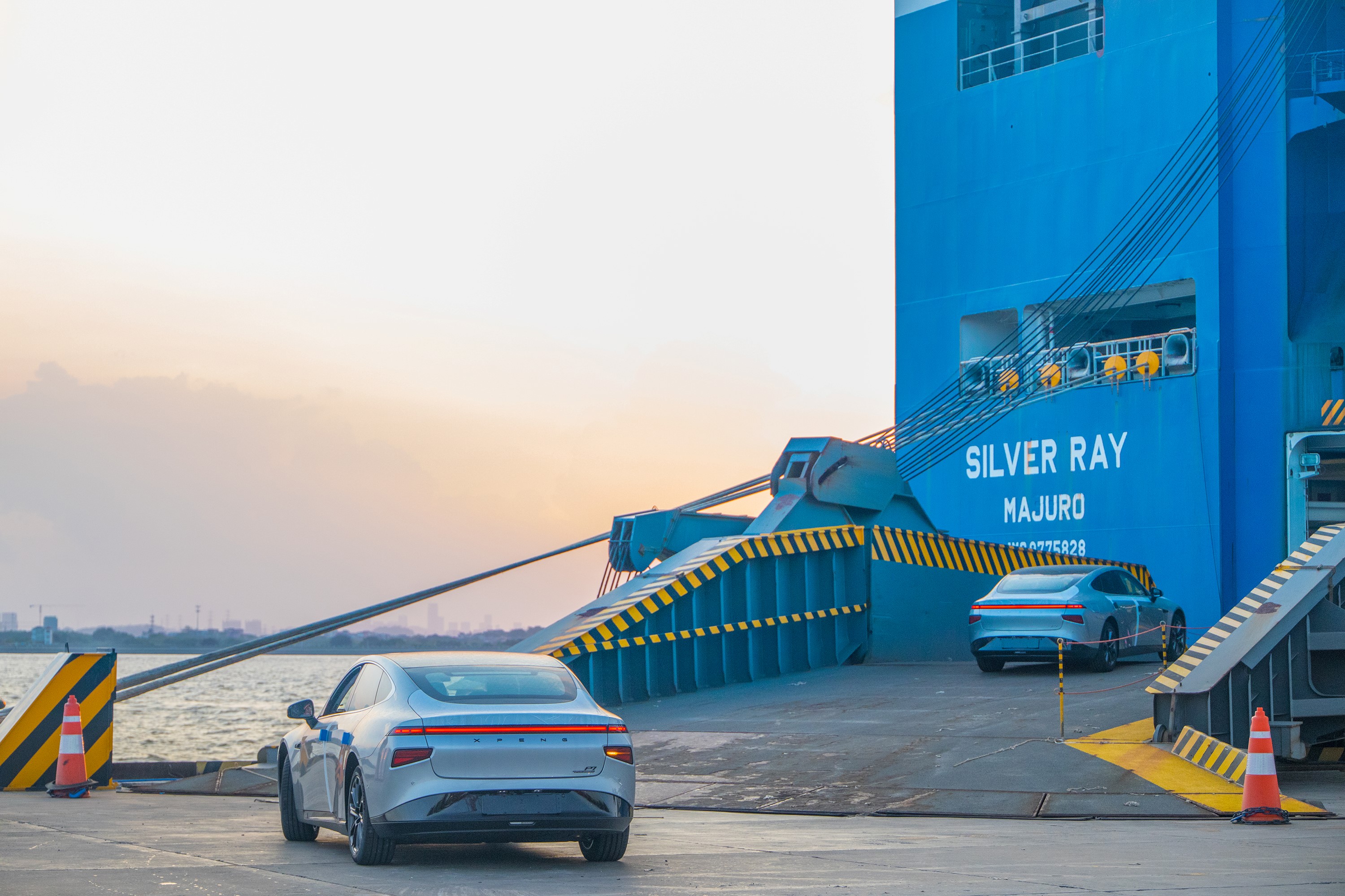 XPeng electric cars being loaded onto the Silver Ray ship at a port during sunset. The image shows vehicles driving up the ramp onto a large blue cargo vessel named 'SILVER RAY MAJURO', illustrating international automotive shipping and logistics.