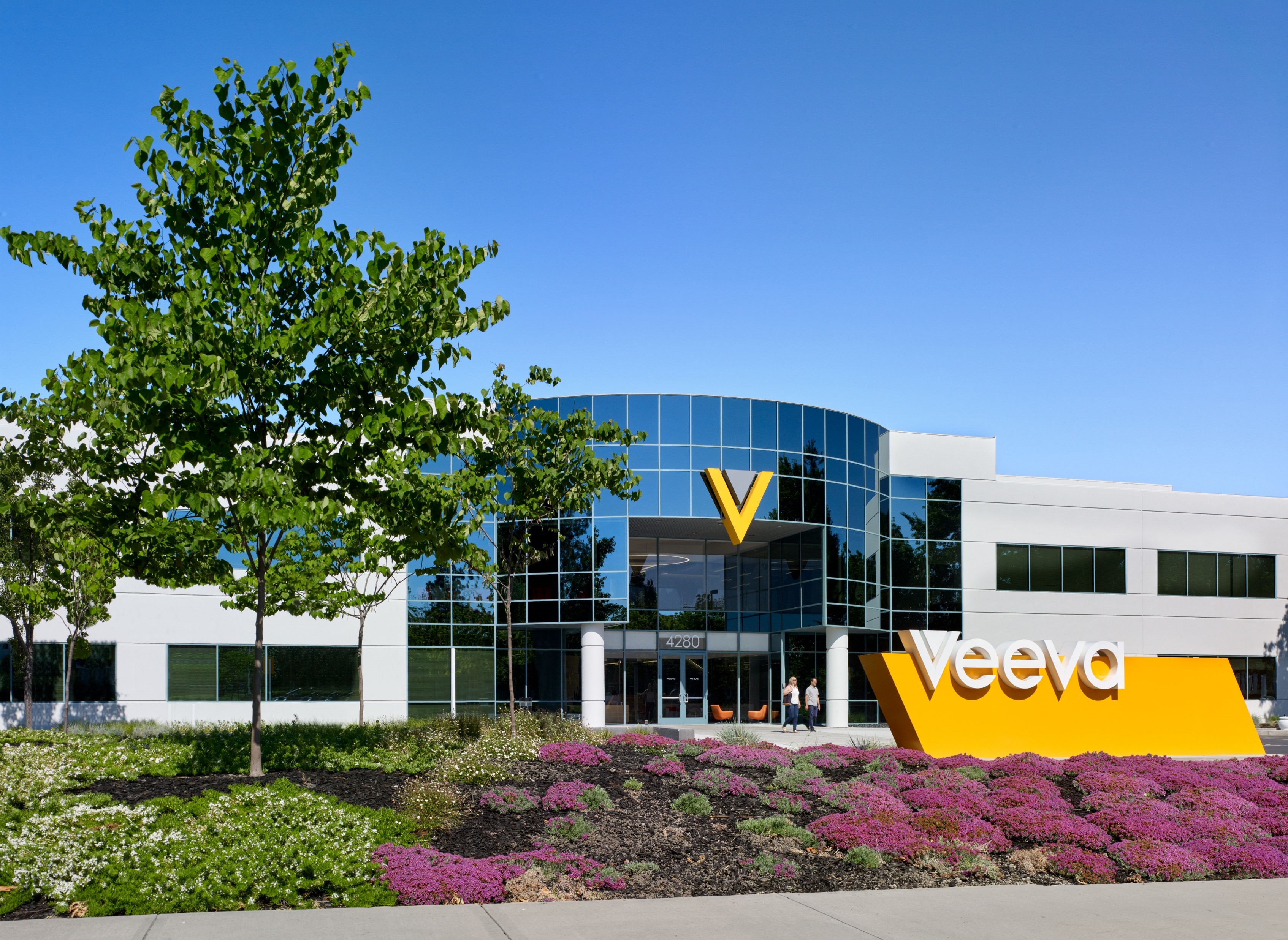 Exterior view of the Veeva headquarters building with glass windows, company signage, landscaping, and a clear blue sky.