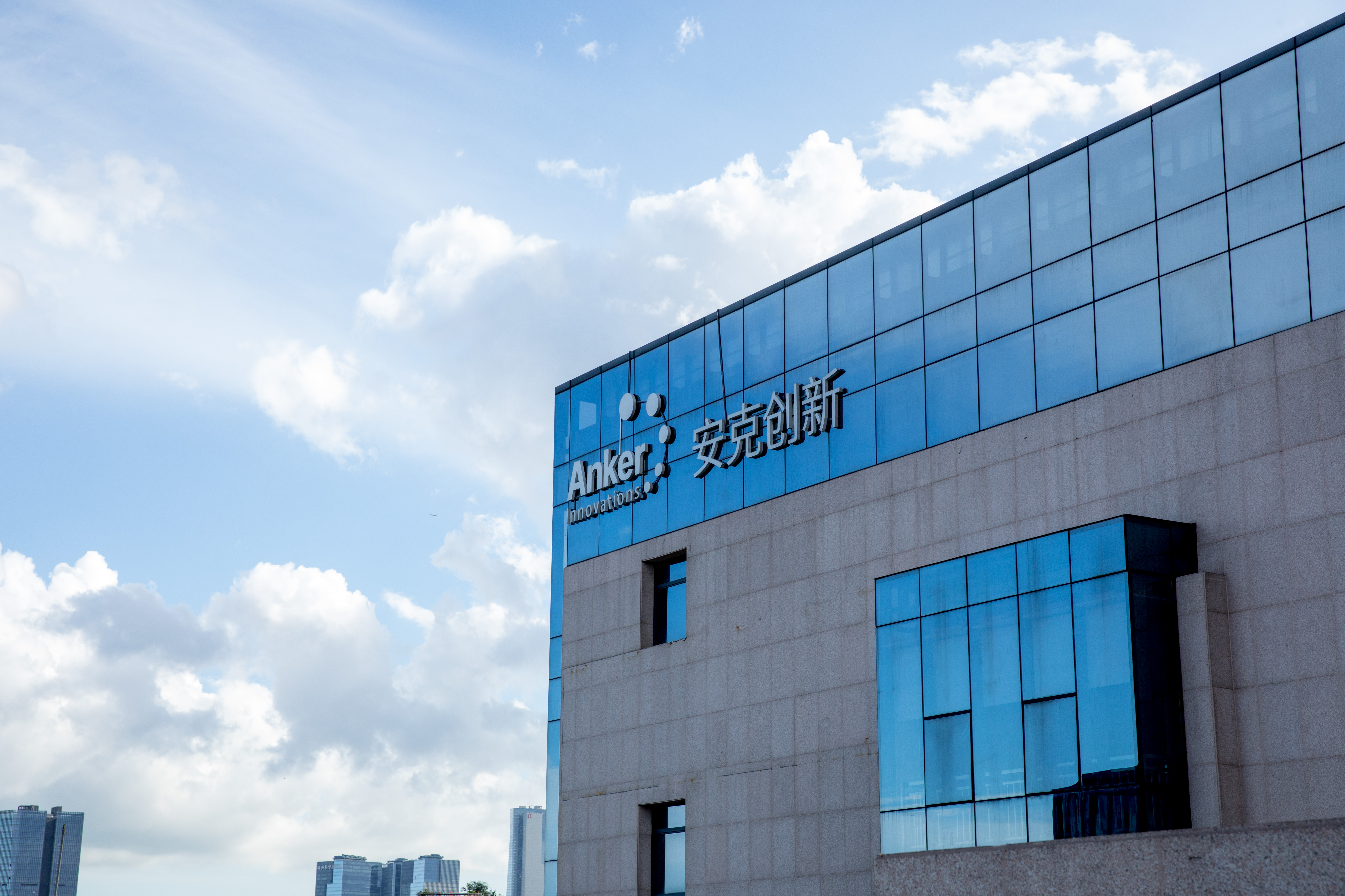 The exterior of the Anker Innovations headquarters building featuring a modern glass facade under a blue sky with clouds. The building has prominent company signage in both English and Chinese.
