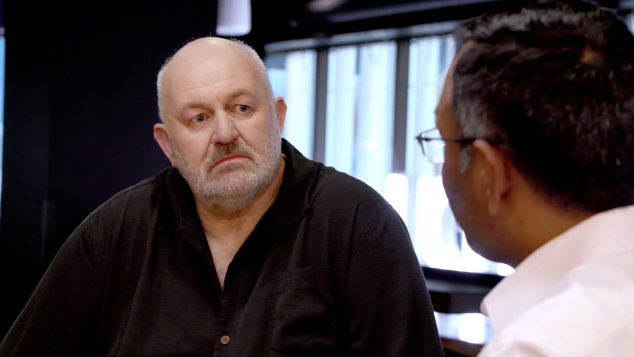 Two men engaged in a genuine conversation in a modern indoor setting. One man is bald and has a contemplative facial expression, while the other man is partially visible and wearing glasses. Light streams in from large windows in the background.