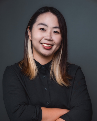 A professional portrait of a smiling woman with long hair, wearing a black shirt, against a dark background.