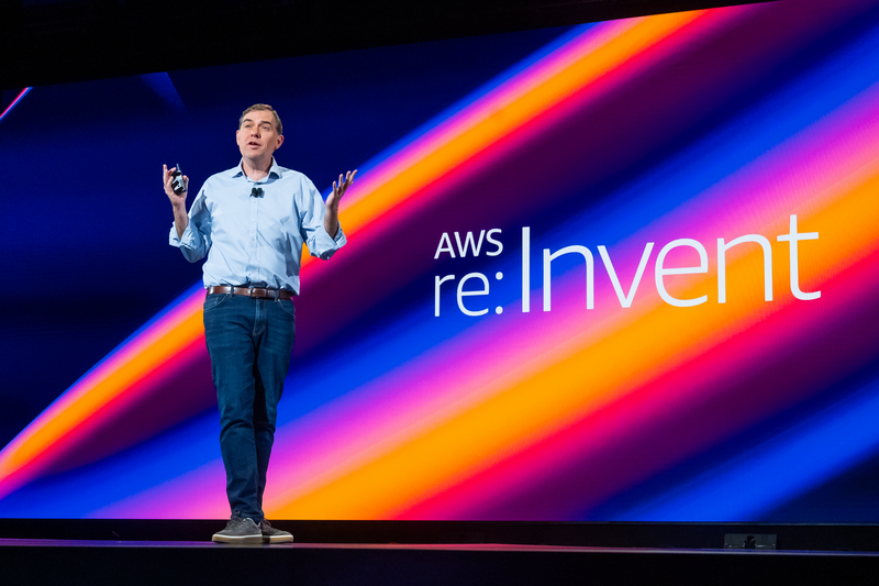 A speaker presenting on stage at AWS re:Invent 2024, with the event logo displayed on a vibrant, colorful background.