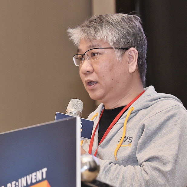 A speaker wearing a grey AWS hoodie and glasses is holding a microphone and speaking at an AWS re:Invent event.
