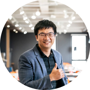 A person in a blue blazer giving a thumbs-up gesture in a modern, well-lit office space.