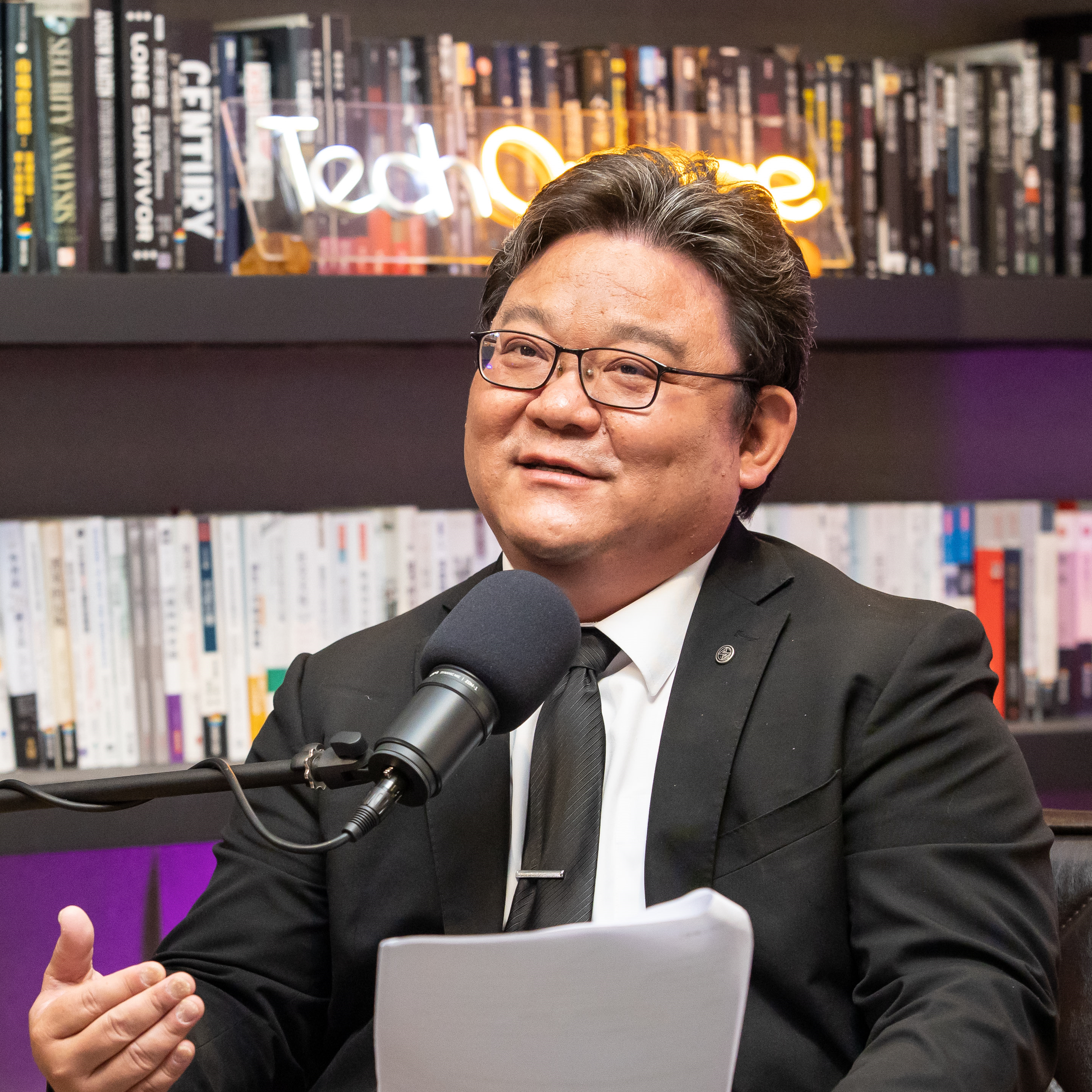 A person in a suit speaking into a microphone, seated in front of a bookshelf with a neon "Tech" sign.