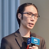 A speaker presenting at an AWS event, holding an AWS-branded microphone. The session is labeled as 303.