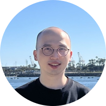 A person standing outdoors near a body of water with cranes and palm trees visible in the background under a clear blue sky.