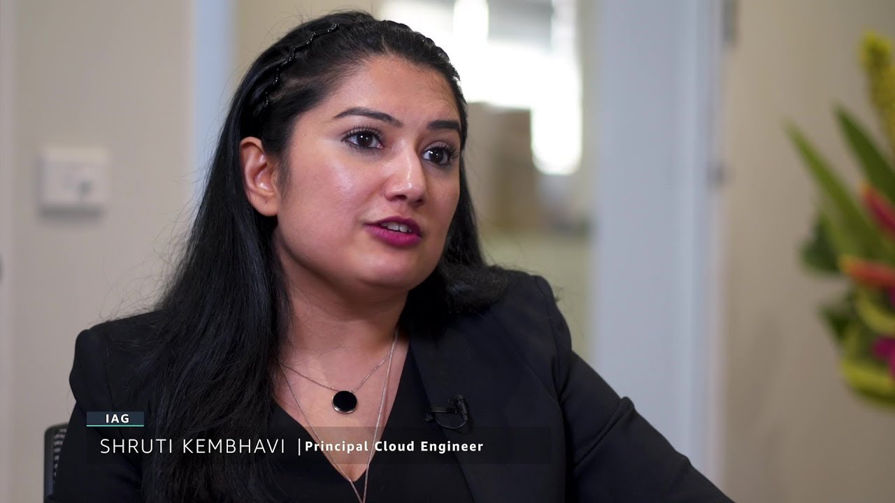 A woman is interviewed in a professional setting. The on-screen text identifies her as Shruti Kembhavi, Principal Cloud Engineer at IAG.