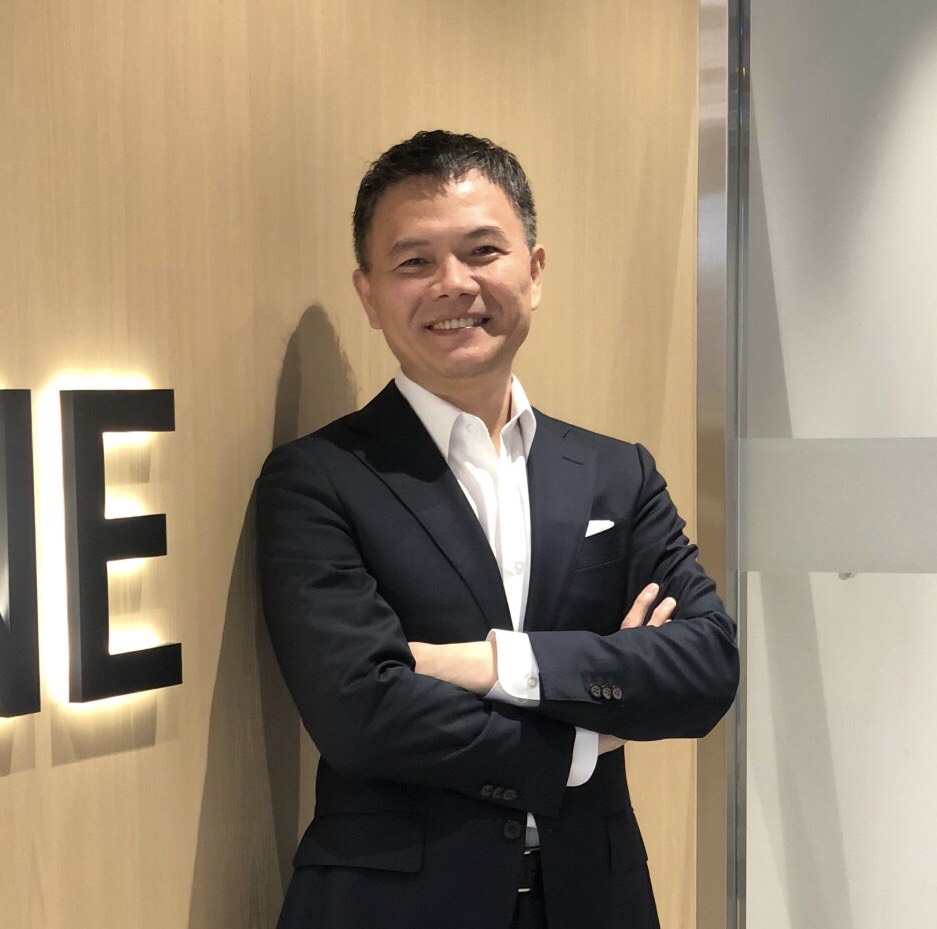 A businessman in a dark suit and white shirt is smiling, standing with arms crossed in an office setting next to a wooden wall.