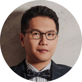 A person wearing a suit, striped shirt, and polka-dot bow tie against a textured wall.
