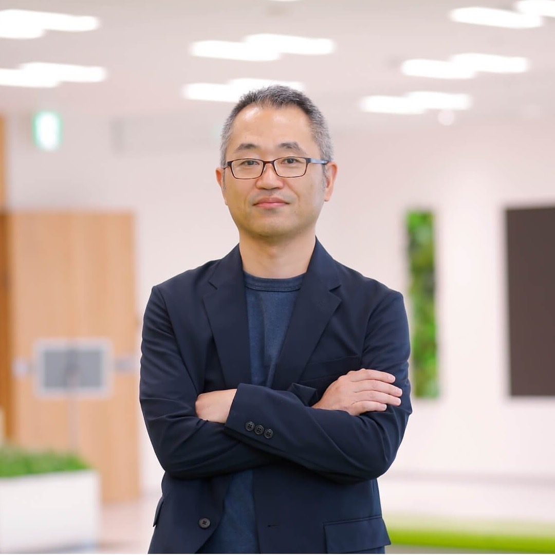 A man wearing business casual attire with his arms crossed, standing in a modern office environment with soft lighting and contemporary decor.