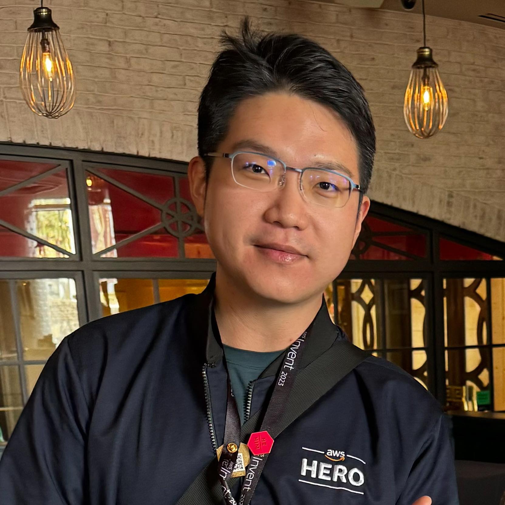 Portrait of an AWS Hero attendee at an AWS event, wearing a jacket with 'AWS HERO' and an event lanyard, pictured indoors with modern lighting.