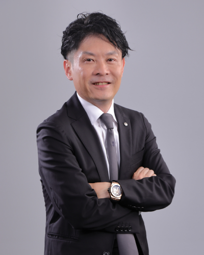A businessman in a black suit and tie standing with arms crossed, smiling, in front of a plain gray background.