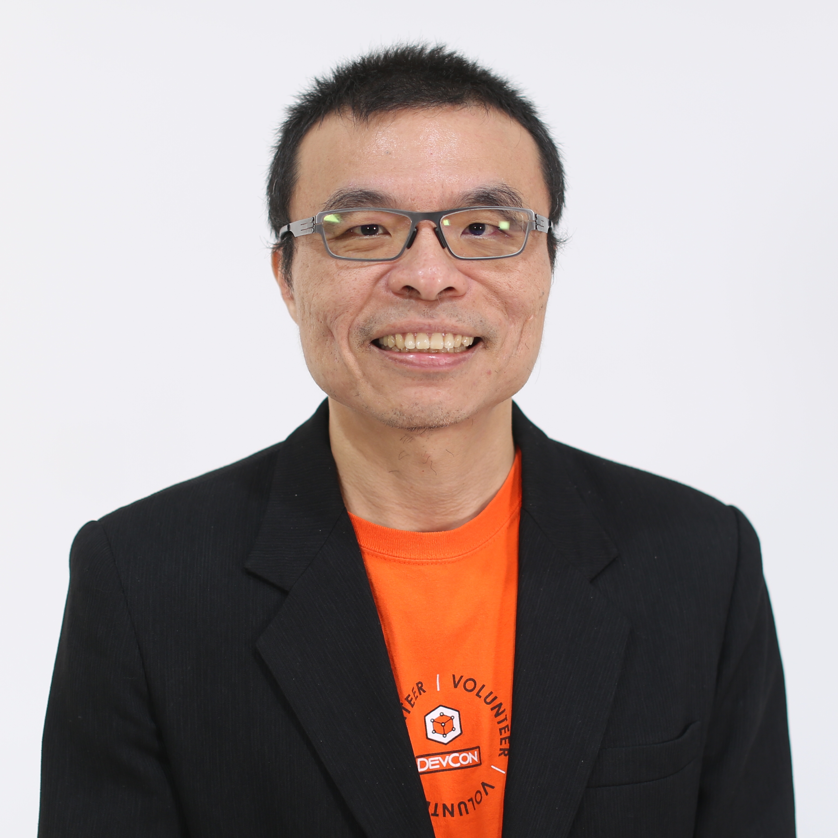A person wearing glasses, an orange t-shirt, and a black blazer, smiling in a studio portrait against a white background.