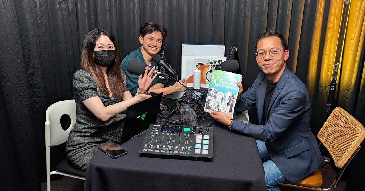 Three people recording a podcast in a studio, seated around a table with microphones, a mixer, a laptop, and a book.