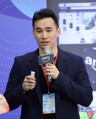 A man holding a microphone and speaking at an AWS event, wearing a suit and event badge, with an AWS-branded microphone and presentation screen in the background.