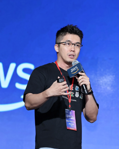 A man speaking at an AWS Summit event, holding a microphone and wearing a black t-shirt and glasses, with an AWS lanyard and badge, standing against a blue background with the AWS logo.