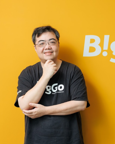A man wearing a black B!GO shirt standing in front of a yellow background, smiling with glasses and posing thoughtfully.