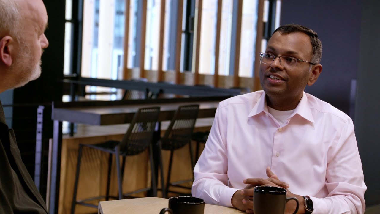 Two individuals engage in a discussion about generative AI at a coffee table, with mugs in front of them, set in a modern office or cafe environment.
