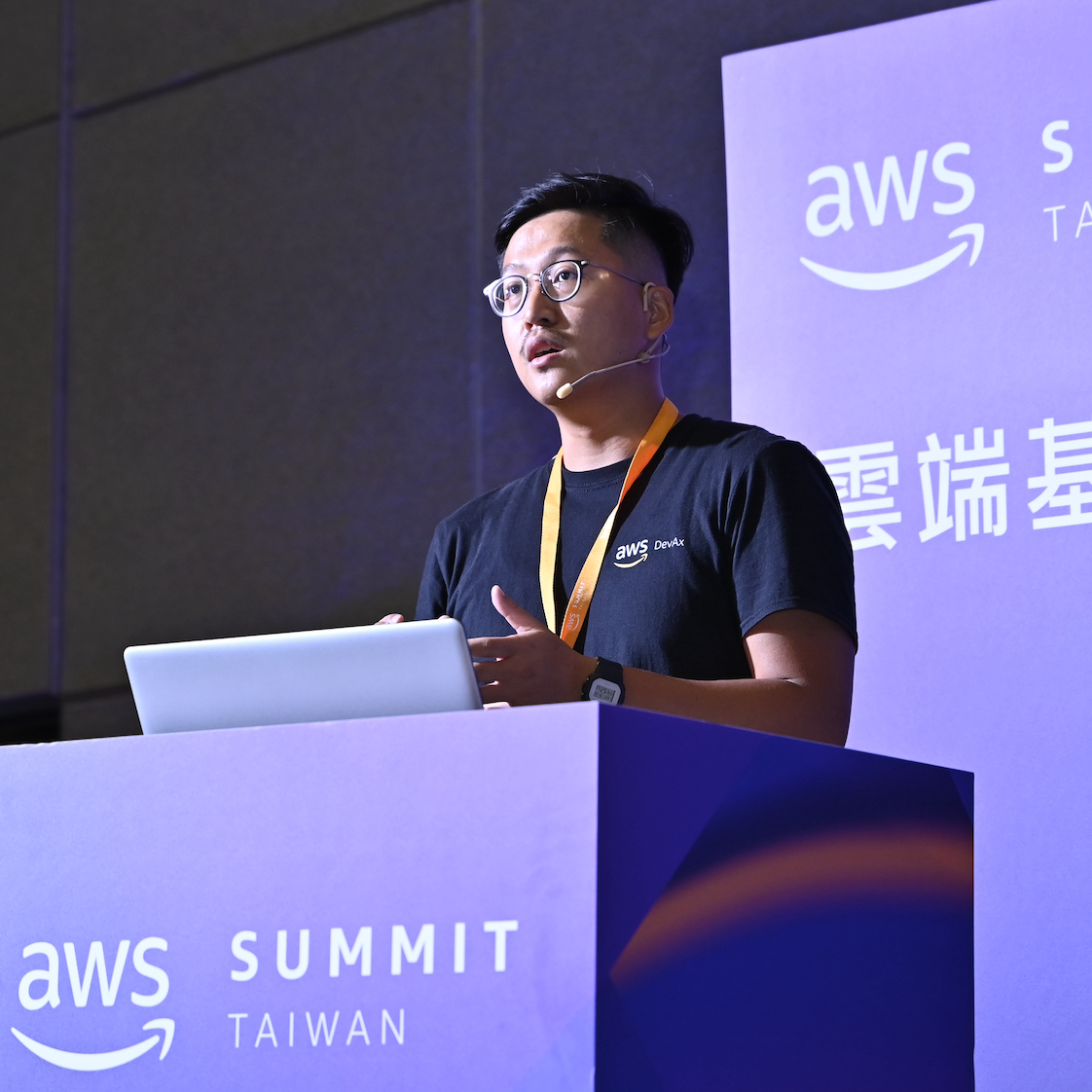A speaker presents at the podium during AWS Summit Taiwan, standing behind a laptop and an event-branded lectern. The individual is wearing an AWS DevAx t-shirt and a headset microphone.
