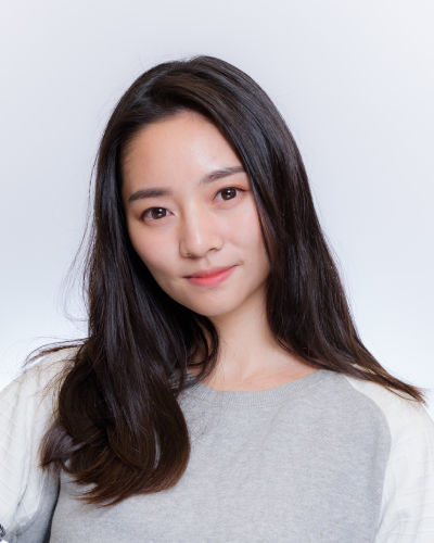 Portrait of a woman with long dark hair, wearing a gray sweater, against a light background.