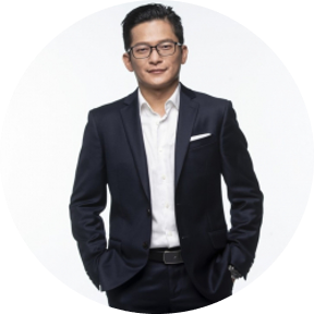 A person wearing a dark suit with a white dress shirt and a pocket square, standing against a plain white background.