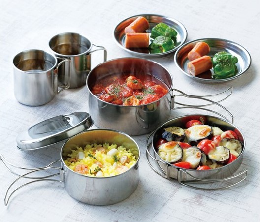 An assortment of cooked food, including vegetables, rice, and meatballs, served in stainless steel camping containers and cups on a white surface.
