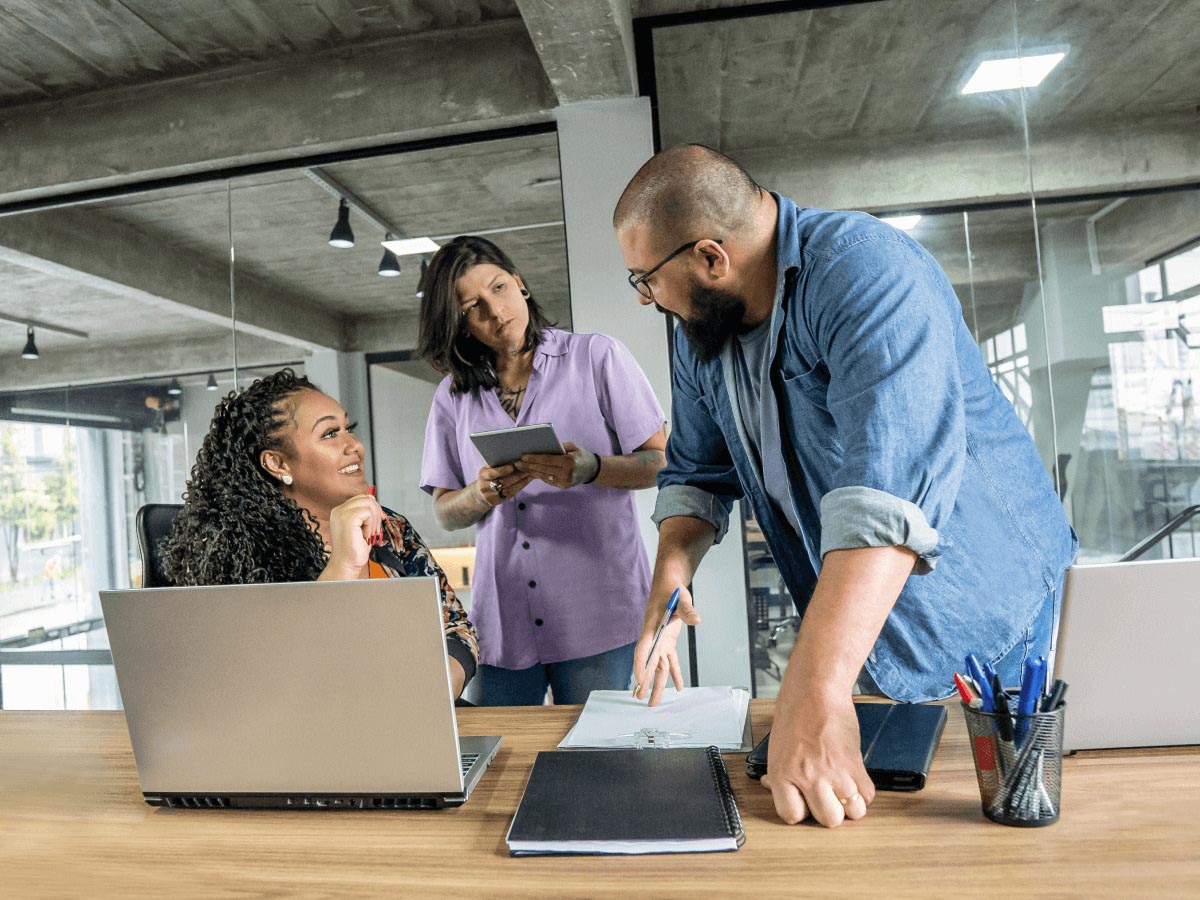 Three people in office, black woman sitting at laptop, tan woman standing, and tan man with beard standing. 
