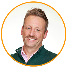 Headshot of a smiling adult man with light hair, wearing a green sweater and pink-collared shirt, shown in a circular frame with an orange border on a white background.