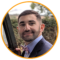 A professional headshot photo of a person wearing a suit and bow tie, standing outdoors with greenery in the background.